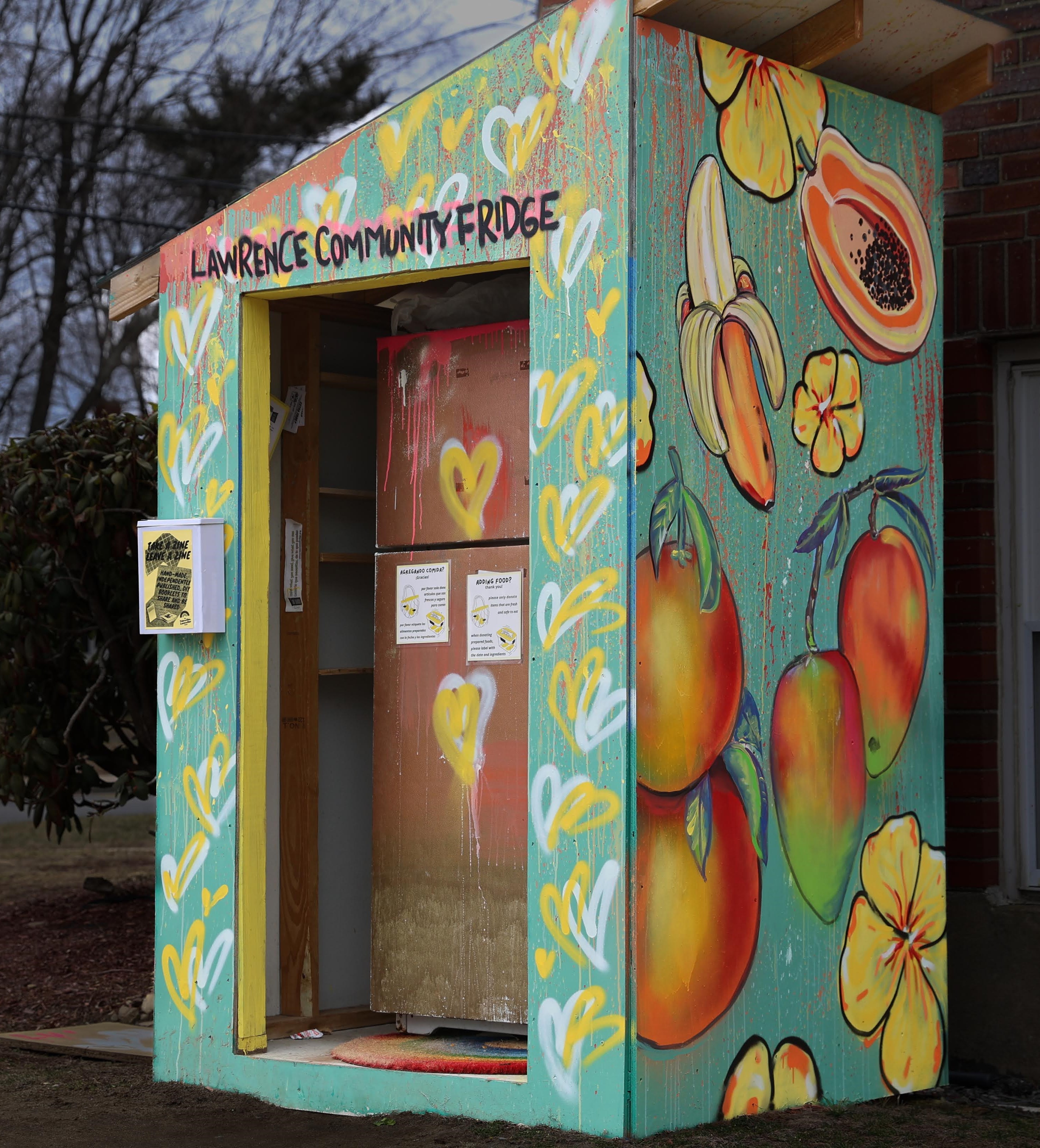 color photo of a refrigerator housed by a shed, both are outside on a grassy area against a brick wall. the shed and fridge are spraypainted many colors. text above the doorway reads 'lawrence community fridge'